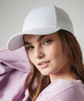 A person with fair skin and long brown hair is wearing a white Beechfield BC648 baseball cap and a pastel pink sweatshirt. They have an expression of calm confidence and are posed with one arm bent, resting near their head, against a neutral background.