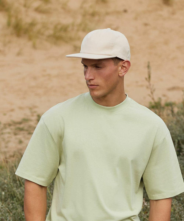 A person with short hair wears a light green oversized T-shirt and a cream-colored Beechfield BC64N cap, looking down and away from the camera. The background features a sandy, grassy outdoor area.