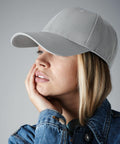 A person with long, blond hair is wearing a light gray Beechfield BC651 baseball cap and a denim jacket. The individual is resting their head on their left hand and looking to the left. The background is plain gray.