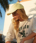 A woman with long blonde hair, wearing a light yellow Beechfield BC653 cap and a white long-sleeve shirt with "Boston" and an eagle logo printed on it, gazes downward. She is outdoors with palm trees and a building partially visible in the background.