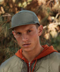 A person wearing a gray Beechfield BC654 baseball cap, orange hoodie, and green jacket stands outdoors with a neutral expression. Trees and foliage are visible in the background.