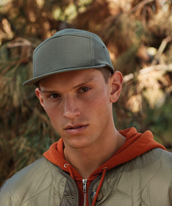 A person wearing a gray Beechfield BC654 baseball cap, orange hoodie, and green jacket stands outdoors with a neutral expression. Trees and foliage are visible in the background.