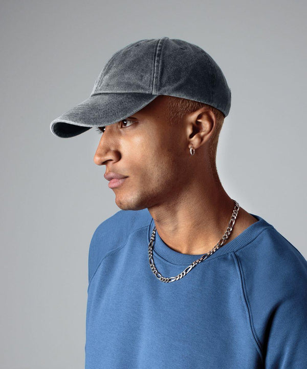 A person with short blonde hair is wearing a gray Beechfield BC655 baseball cap, a blue sweatshirt, a silver necklace, and a small hoop earring. They are facing to the right against a plain gray background.