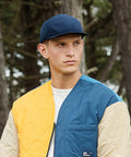 A person wearing a quilted jacket with blue, yellow, and beige patches and a Beechfield BC659 navy blue cap stands outdoors against a background of trees. The individual looks slightly to the side with a neutral expression.