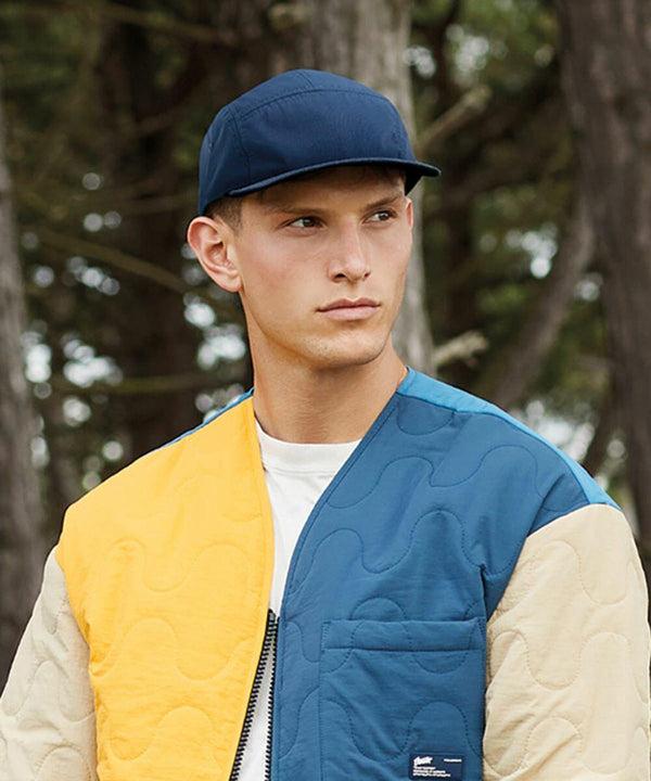A person wearing a quilted jacket with blue, yellow, and beige patches and a Beechfield BC659 navy blue cap stands outdoors against a background of trees. The individual looks slightly to the side with a neutral expression.