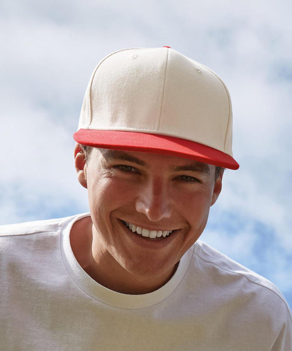 A smiling person wears a white Beechfield Contrast flat peak 6-panel cap with a red brim and a white T-shirt, standing outdoors under a partly cloudy sky.