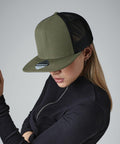 A person wearing a dark olive green Beechfield BC664 trucker cap with a black mesh back, styled with silver hoop earrings and a black zippered top. The individual's head is slightly tilted down, obscuring their face partially from view. The background is plain grey.