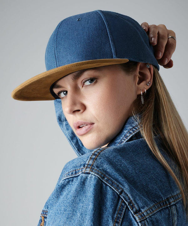 A person with long hair wearing a blue and tan Beechfield BC668 cap and a blue denim jacket is posing with a direct gaze. The Beechfield BC668 cap is tilted slightly, and the person's head is turned to the side. Multiple earrings are visible in their left ear. The background is plain gray.