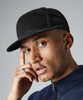 A person with short hair wearing a black Beechfield BC670 cap and a hoop earring is posing with a thoughtful expression. They are resting one finger on the side of their face and dressed in a dark zip-up jacket. The background is plain and grey.