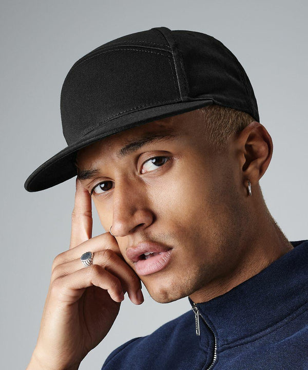 A person with short hair wearing a black Beechfield BC670 cap and a hoop earring is posing with a thoughtful expression. They are resting one finger on the side of their face and dressed in a dark zip-up jacket. The background is plain and grey.