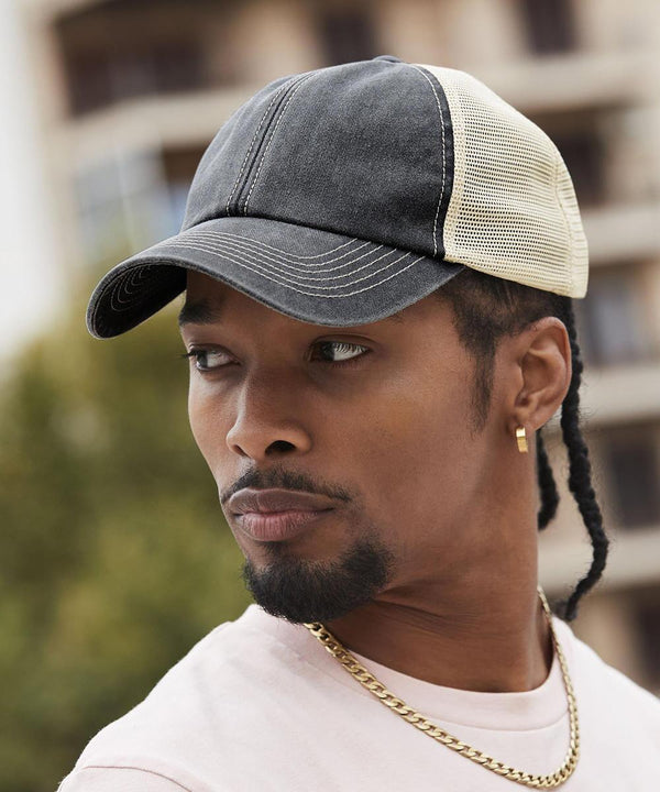 A person wearing a Beechfield 6-panel vintage trucker cap in black and beige mesh, a gold chain necklace, and a light shirt looks to the side outdoors with blurred buildings and greenery in the background.