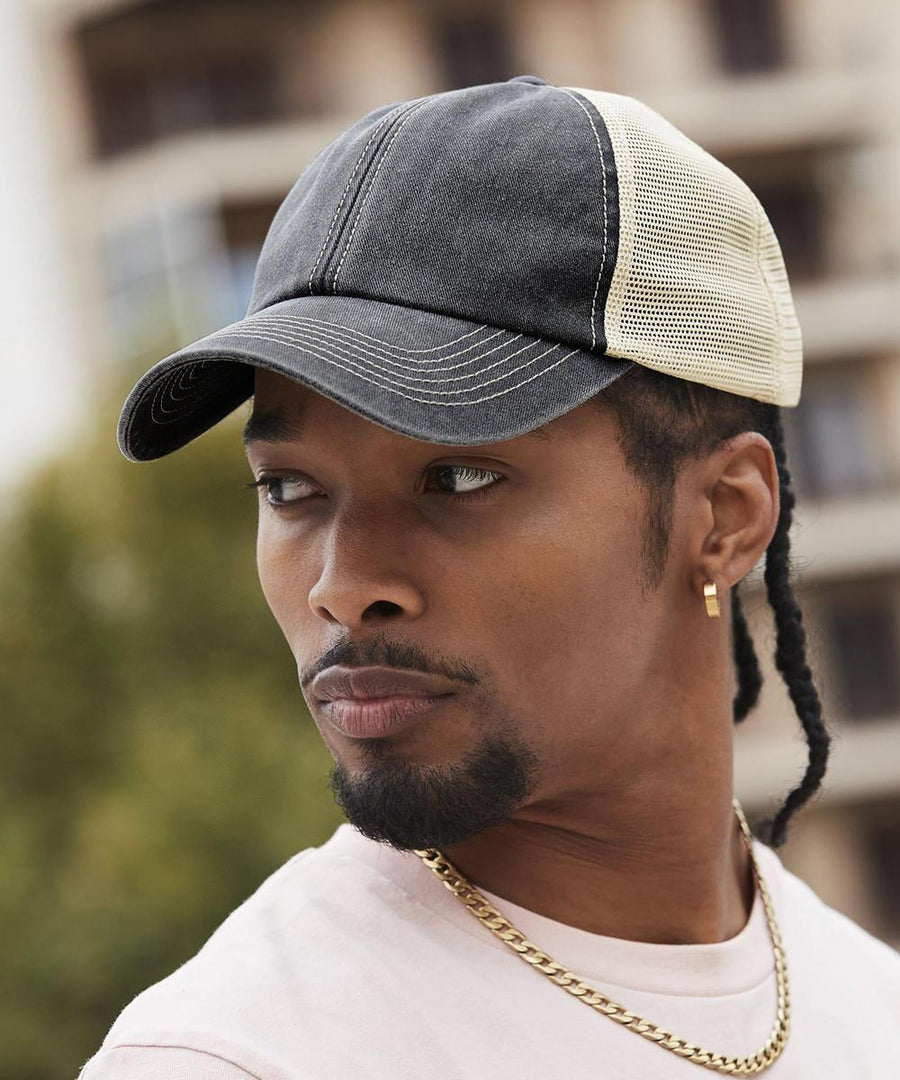 A person wearing a Beechfield 6-panel vintage trucker cap in black and beige mesh, a gold chain necklace, and a light shirt looks to the side outdoors with blurred buildings and greenery in the background.