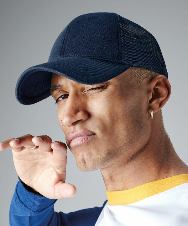 A person wearing a blue Beechfield BC678 cap and a white shirt with blue sleeves and a yellow collar winks while holding a hand up near their face. The person has an earring in the left ear and is posing against a plain grey background.