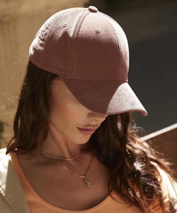 A person with long brown hair wears a Beechfield EarthAware organic cord baseball cap, a peach vest, layered necklaces, and looks down against a softly lit, blurred background.
