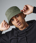 A person with a serious expression poses while holding the brim of their green Beechfield BC686 bucket hat with both hands. They are wearing a black hoodie and have small hoop earrings. The background is plain gray.