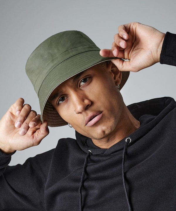 A person with a serious expression poses while holding the brim of their green Beechfield BC686 bucket hat with both hands. They are wearing a black hoodie and have small hoop earrings. The background is plain gray.