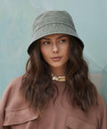 A woman with wavy brown hair wears the Beechfield Vintage bucket hat in green and a brown top, standing before a teal background and gazing to the side with a neutral expression.