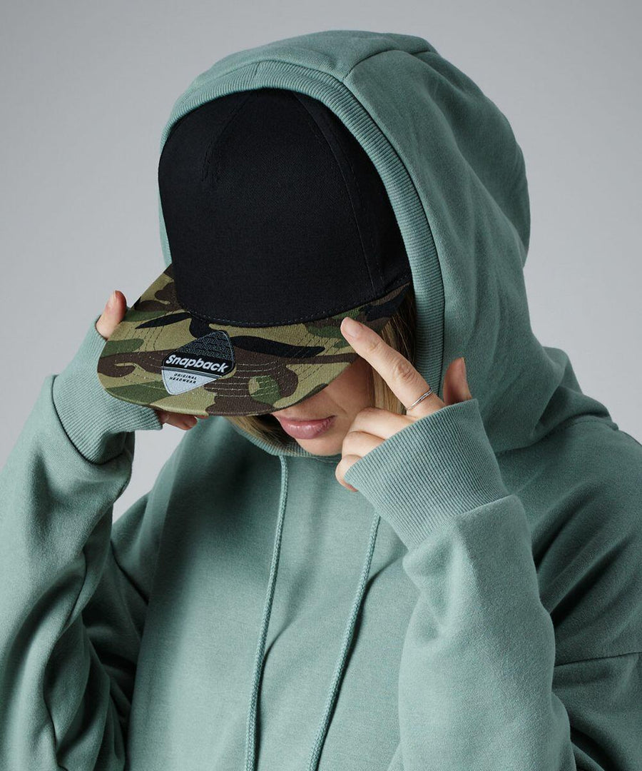 Person in a teal hoodie with the hood up, wearing a Beechfield BC691 black and camouflage snapback cap, tilting their head down, and adjusting the cap with both hands. The background is plain gray.