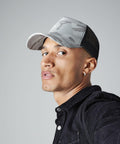 A person with light brown skin and short, blonde hair is wearing a Beechfield BC694 camouflage baseball cap and a black jacket. They have an earring in their left ear. The background is plain gray. They are looking directly at the camera.