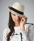 A person with long dark hair is wearing a white and black tie-dye shirt and a Beechfield BC720 straw fedora with a black band. They are slightly tilting the hat with one hand and smiling softly, while partly concealing one eye. The background is plain grey.