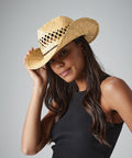 A person with long, dark hair wearing a sleeveless black top and a woven straw Beechfield BC735 cowboy hat. They are tilting the hat with one hand, and smiling slightly, standing against a plain gray background.
