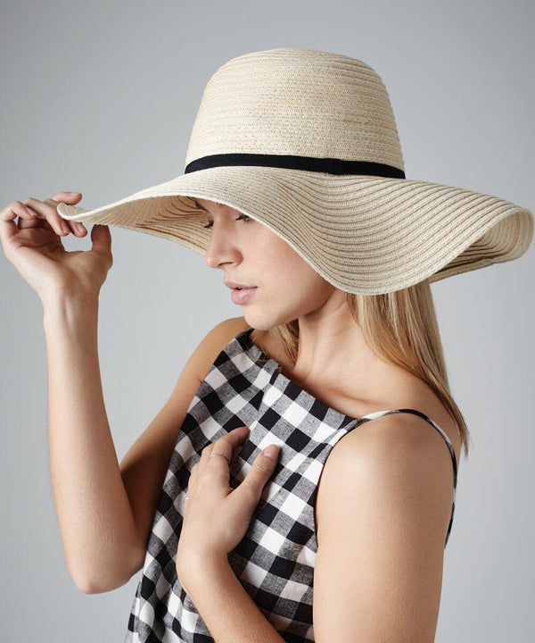 A person with long, light hair wearing a wide-brimmed straw hat with a black ribbon and a black and white checkered sleeveless top. They are tilting the brim of the Beechfield BC740 with one hand and their other hand is resting on their chest. The background is plain gray.
