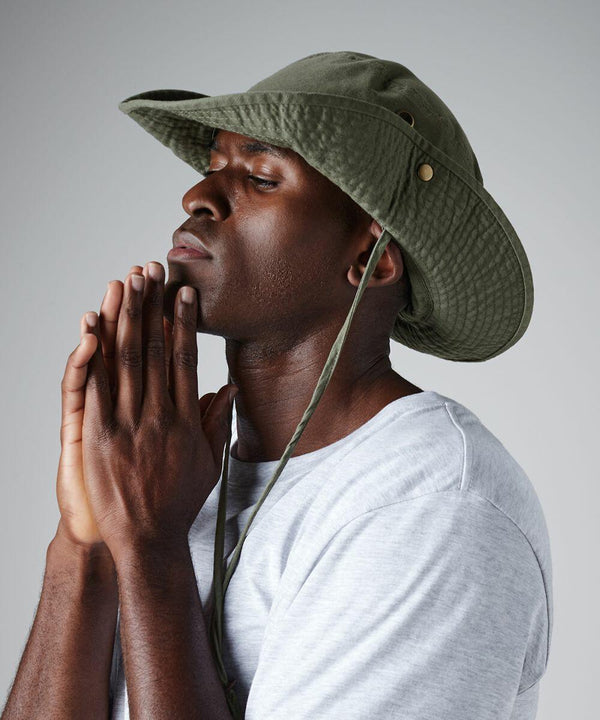 A man wearing an olive green Beechfield BC789 wide-brimmed hat with chin strap clasps his hands together near his face. He is dressed in a light gray t-shirt and appears to be deep in thought or contemplative. The background is plain and light-colored.