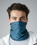 A person with short, wavy hair is wearing a blue and black speckled Beechfield BC901 neck gaiter covering their nose and mouth. They are dressed in a light-colored jacket and standing against a plain gray background.