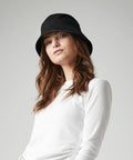 A woman wearing a black Beechfield BC90N and a long-sleeve white shirt is posing against a plain background. Her brown hair is loosely wavy and she has a neutral expression on her face.