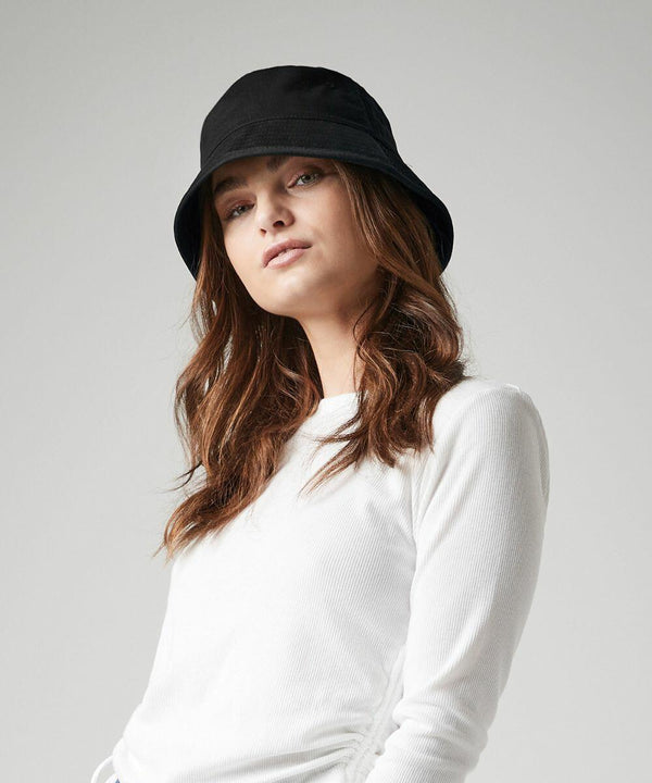 A woman wearing a black Beechfield BC90N and a long-sleeve white shirt is posing against a plain background. Her brown hair is loosely wavy and she has a neutral expression on her face.