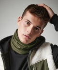 A young man with short brown hair touches his head with one hand while looking into the camera with a serious expression. He is wearing a green Beechfield BC916 neck warmer, a black shirt, and a jacket with a fleece-lined hood. The background is plain and gray.