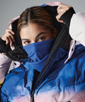 A person with brown hair is wearing a blue and pink gradient puffer jacket and a Beechfield BC920 blue scarf covering their nose and mouth. They are looking directly at the camera while lifting the hood of their jacket.