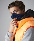 A person with short, curly hair is wearing an orange puffer vest over a grey hoodie. They have a dark blue Beechfield BC930 scarf covering their nose and mouth, holding it in place with one hand. The background is a plain grey.