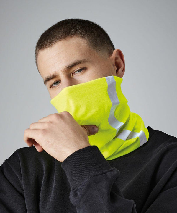 A person with short hair is wearing a fluorescent yellow Beechfield BC950 neck gaiter over their nose and mouth. They are dressed in a black top and are looking at the camera with their head slightly tilted. The background is plain and grey.