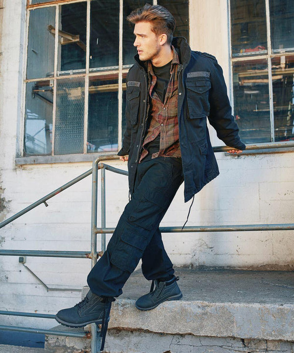A person with styled hair stands on a concrete structure's edge beside a metal railing. They wear a dark jacket over a plaid shirt, dark cargo pants, and matching boots. The backdrop features a weathered wall with industrial-style windows, highlighting the stylish touch of the BD003 from Build Your Brandit.