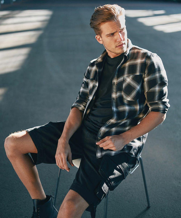 A man with light hair sits on a stool in a sunlit area wearing a black checkered flannel shirt over a black shirt, black cargo shorts, and BD201 boots by Build Your Brandit. He gazes off to the side, with shadows and light creating patterns on the floor and background.