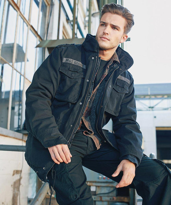 A man with short hair is sitting on a metal railing in front of an industrial building. He is wearing a Build Your Brandit BD301 jacket, buttoned-up and layered over a flannel shirt. The background includes glass windows and metal structures.