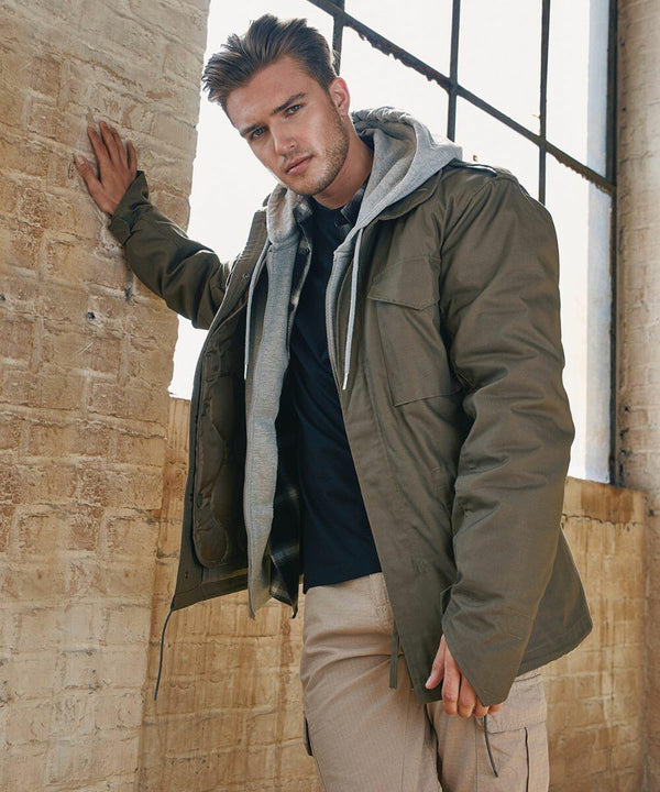 A man wearing a green jacket layered over a gray hoodie leans against a tall, sunlit brick wall, looking at the camera. His left hand is resting on the wall while his right hand is down at his side. He is dressed in casual light brown Build Your Brandit BD308 pants.