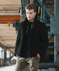 A man with styled hair is standing in a partially industrial indoor setting. He is wearing a black jacket over a dark shirt and khaki pants, with his hands in his jacket pockets. He is looking directly at the camera with a neutral expression, showcasing the BD316 by Build Your Brandit.