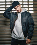 A man wearing a black beanie, dark bomber jacket, and grey BD349 by Build Your Brandit stands in front of a metal shutter, tilting his head slightly while holding his beanie with one hand. The background is lit by light coming through the shutter slats.
