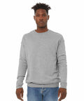 A person stands against a plain white background, wearing a Bella Canvas BE045 light gray long-sleeve sweatshirt and blue jeans. They have short, styled hair and a neutral facial expression.