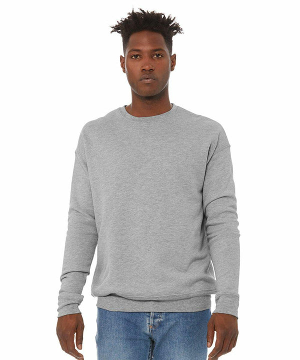 A person stands against a plain white background, wearing a Bella Canvas BE045 light gray long-sleeve sweatshirt and blue jeans. They have short, styled hair and a neutral facial expression.