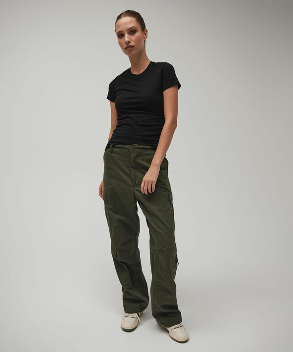 A woman wearing a black Bella Canvas BE076 T-shirt and olive green cargo pants stands against a plain background. She has her hair tied back and is looking directly at the camera. She is also wearing white sneakers.