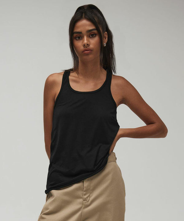A woman with long dark hair wears a Bella Canvas BE089 black tank top and beige pants. She stands confidently, one hand on her hip and the other relaxed at her side, against a plain gray background.