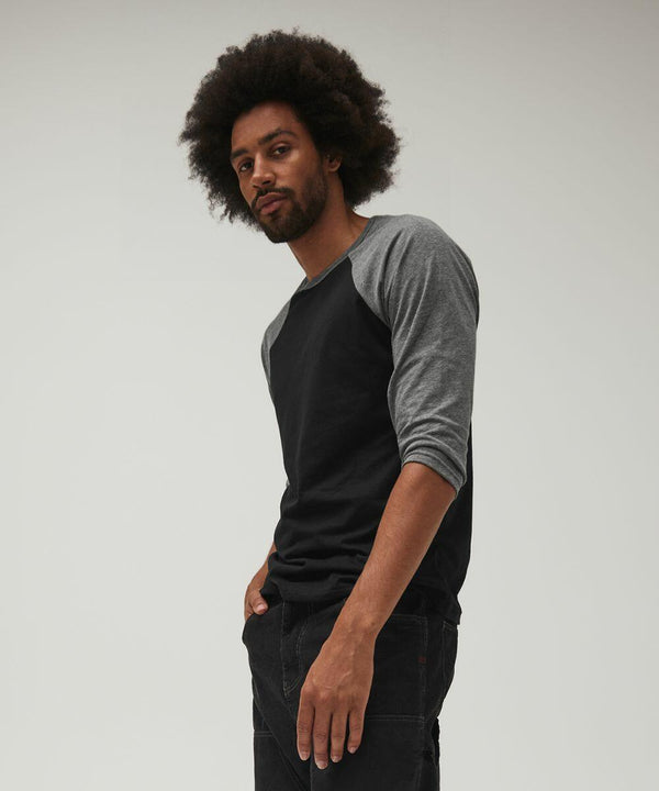 A person with curly hair and a beard stands facing slightly sideways, looking towards the camera. They are wearing a black and gray long-sleeved Bella Canvas BE100 shirt and dark pants. Their right hand is in the pocket of their pants. The background is plain and light-colored.