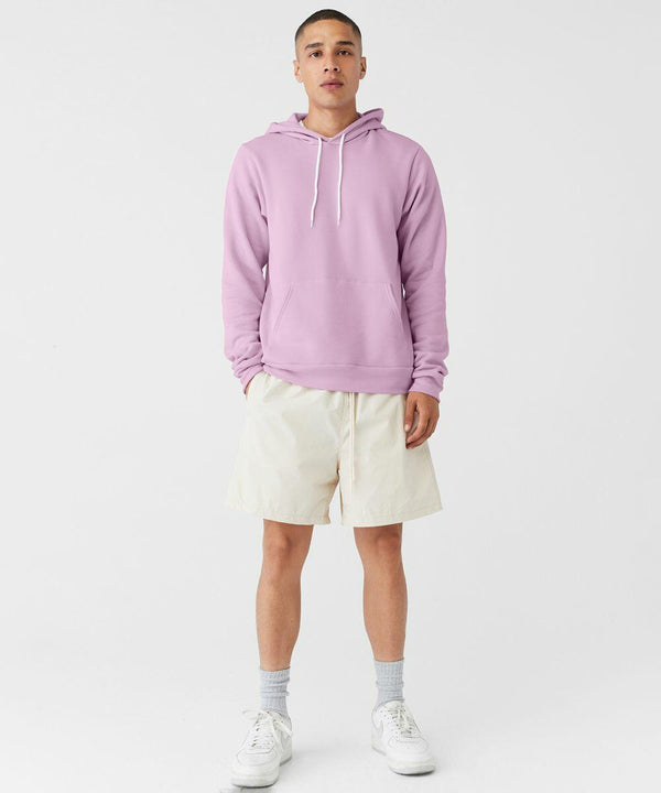 A person stands facing forward against a plain white background. They are wearing a light purple Bella Canvas BE105 hoodie with a front pocket and white drawstrings, light-colored shorts, gray socks, and white sneakers. The person has a neutral expression and their hands are in their pockets.