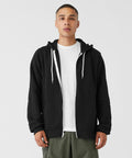 A person stands facing the camera wearing a Bella Canvas BE106 black zip-up hoodie with white drawstrings and a white T-shirt underneath. They also wear olive green pants. The background is plain and white.