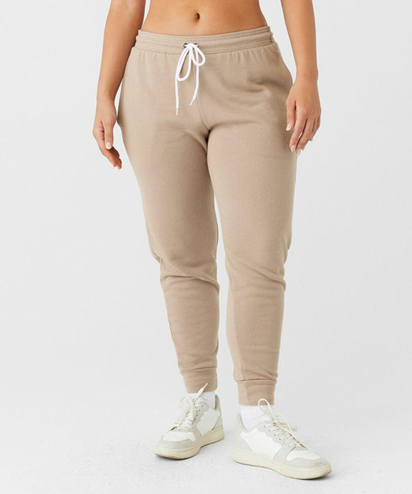 A person wearing Bella Canvas BE126 beige jogger pants with a white drawstring and white sneakers stands against a plain white background. The pants have a relaxed fit and are paired with casual athletic shoes. The midsection of the person is visible, while the upper body is not shown.
