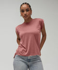 A person with braided hair stands confidently, wearing a rose-colored Bella Canvas BE128 sleeveless top and light blue jeans. They have hoop earrings and a neutral expression, with their hands resting by their sides. The background is plain and light gray.