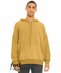 A young person wearing a mustard-colored Bella Canvas BE130 hoodie with a front pocket and drawstrings stands against a white background. They have a neutral expression and their hands are by their sides. The text "fwd FASHION" is displayed on the lower part of the image.